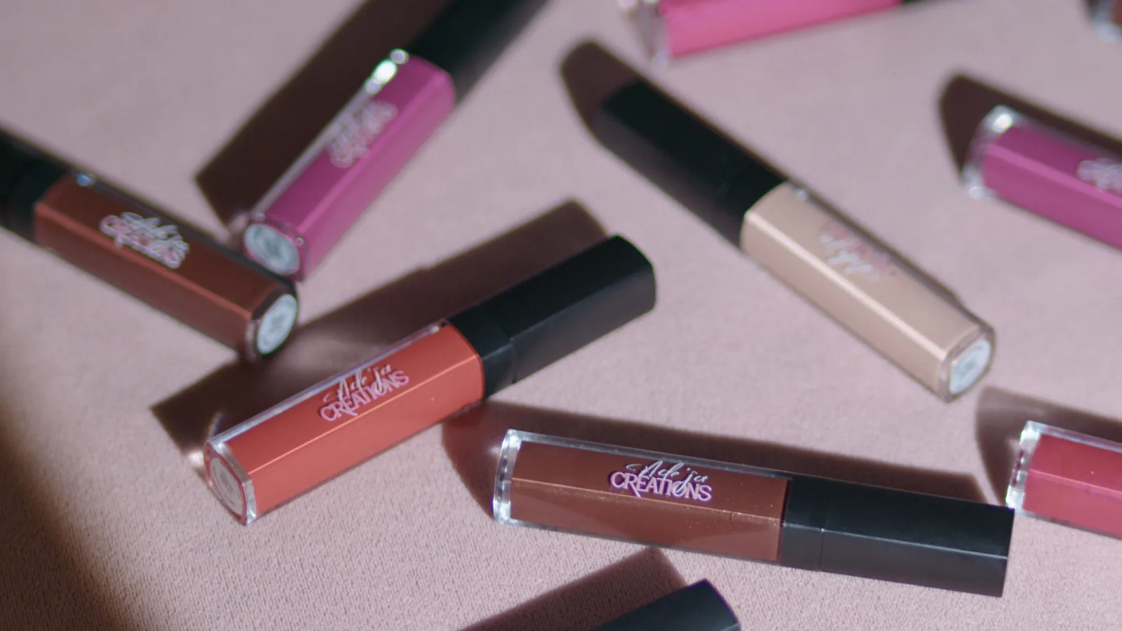 Lip gloss tubes scattered across pink paper in hard afternoon light.