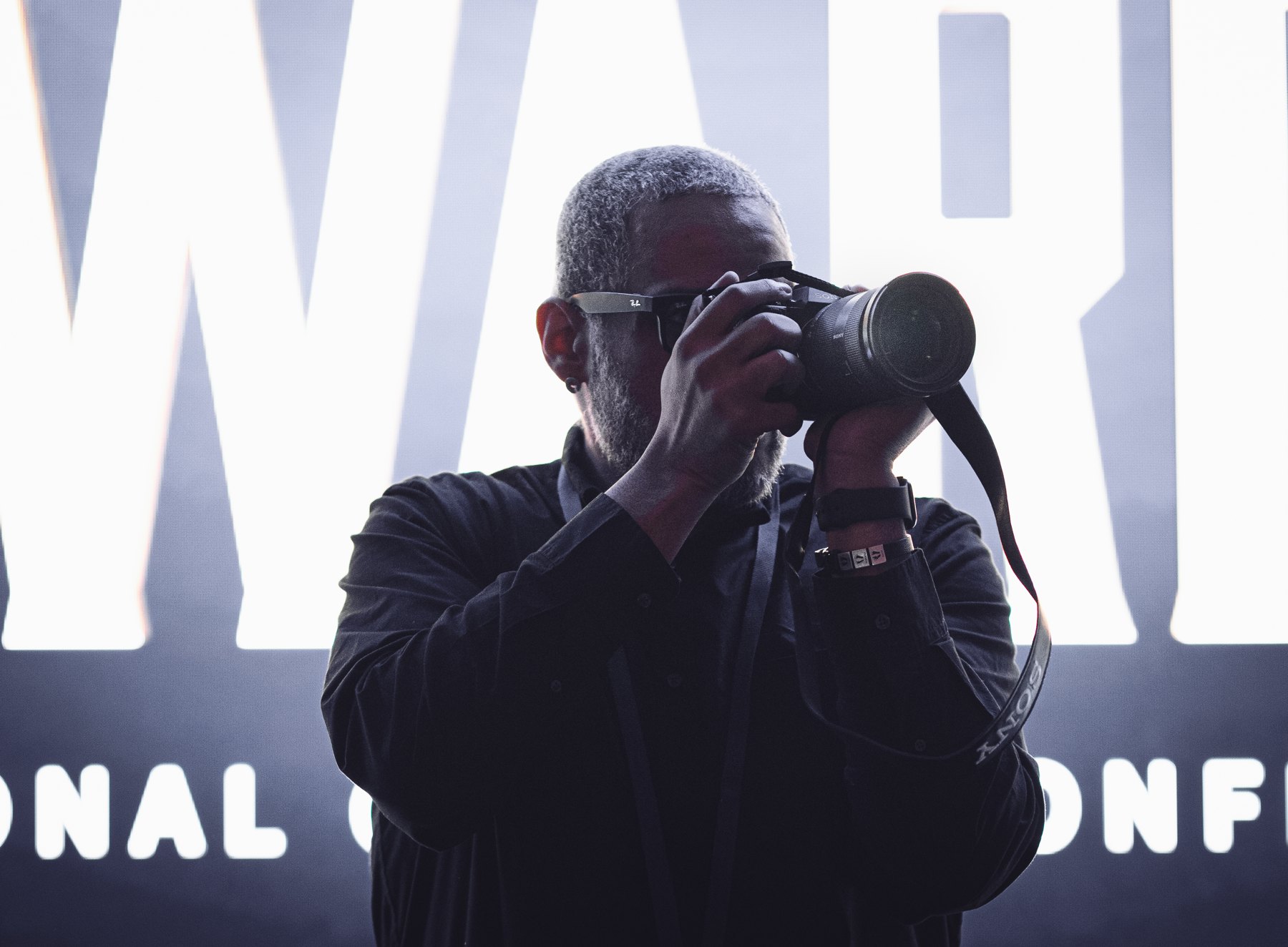 Falú at work, camera to his eye, in front of a lit backdrop.