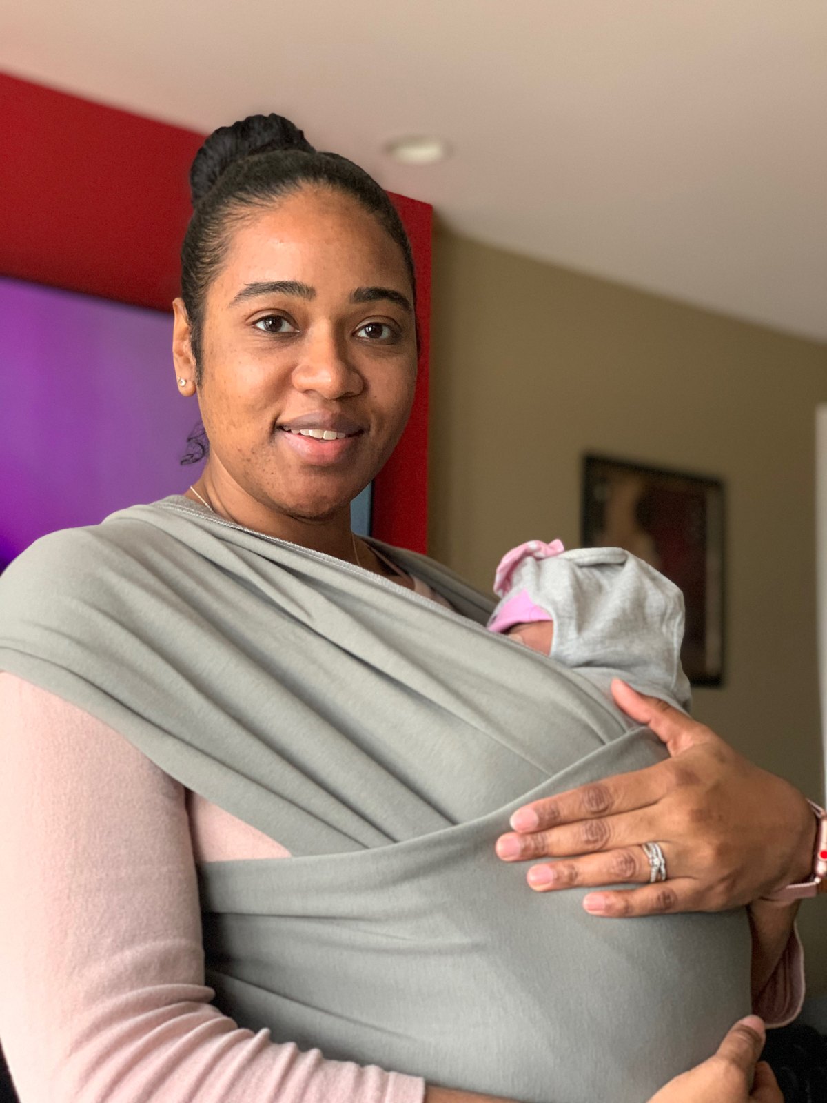 Indira smiling, carrying baby Havana in a grey wrap at home.